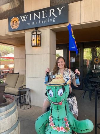 Woman holding two wine bottles at a winery wine-tasting entrance, posing behind a large playful cactus mascot wearing a straw hat and flower lei on a sunny outdoor patio with seating.