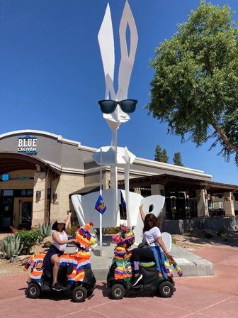 Two people on bright piñata-covered mobility scooters pose in front of a giant white rabbit sculpture wearing sunglasses at a sunny outdoor plaza with trees and storefronts.