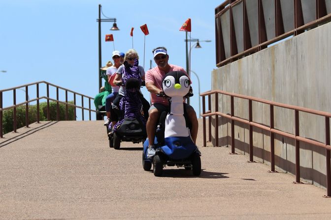 Group of adults riding novelty animal-themed mobility scooters (penguin, zebra) down a sunlit beachside promenade ramp with railings and lamp posts