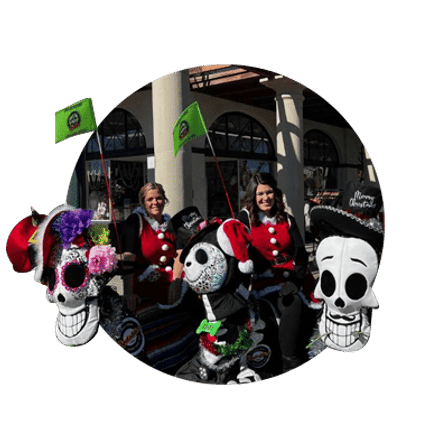 Festive outdoor market stall featuring colorful Day of the Dead–style skull and skeleton decorations with two vendors in red jackets under arched storefronts.