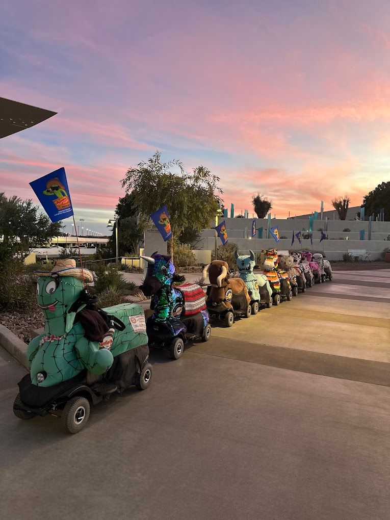 Flag-topped, character-themed ride-on mobility scooters lined up on a modern outdoor plaza at sunset, colorful costumes and palm trees beneath a pink sky.
