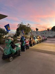 Flag-topped, character-themed ride-on mobility scooters lined up on a modern outdoor plaza at sunset, colorful costumes and palm trees beneath a pink sky.