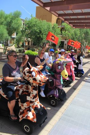 Group of adults riding colorful animal- and unicorn-shaped mobility scooters, waving orange flags during a sunny outdoor plaza parade with desert landscaping.