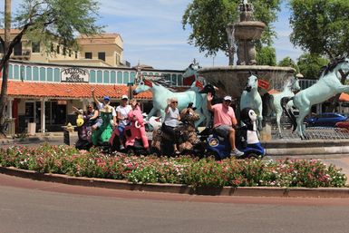 Group of people on colorful animal-shaped mobility scooters waving in front of a horse fountain and flower-filled roundabout in an Arizona shopping plaza