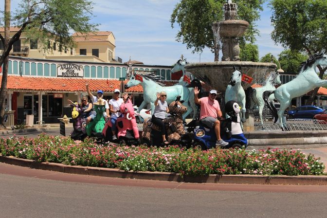 Group of people on colorful animal-shaped mobility scooters waving in front of a horse fountain and flower-filled roundabout in an Arizona shopping plaza