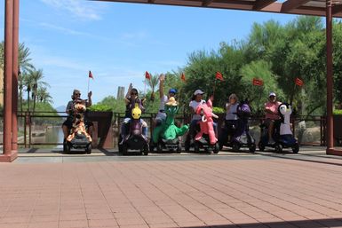 Group of people on colorful animal-shaped mobility scooters lined up on a sunny waterfront promenade with palm trees and desert greenery, waving under a shaded pavilion