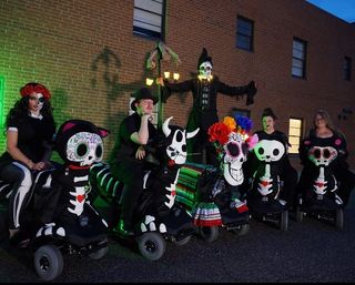 Group of Day of the Dead revelers in skeleton costumes and painted skull masks riding decorated mobility scooters in front of a brick building at night, with colorful paper flowers and a tall illuminated skeleton figure in the background.