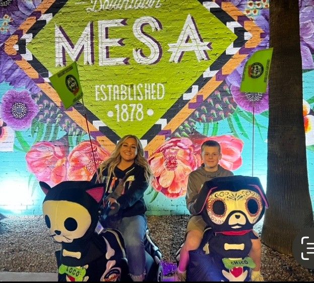 Two smiling riders on colorful skeleton-style kiddie rides in front of a bright Downtown Mesa 'Established 1878' floral mural at night.