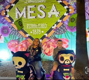 Two smiling riders on colorful skeleton-style kiddie rides in front of a bright Downtown Mesa 'Established 1878' floral mural at night.