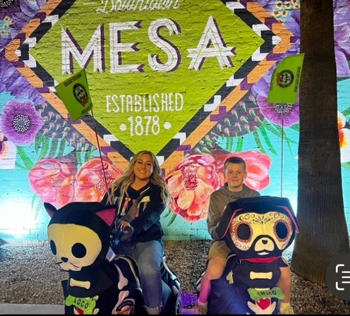 Two smiling riders on colorful skeleton-style kiddie rides in front of a bright Downtown Mesa 'Established 1878' floral mural at night.