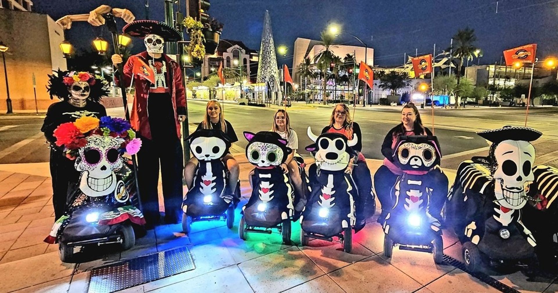 Group of people in Day of the Dead skeleton costumes on decorated mobility scooters lined up on a downtown city plaza at night, with colorful LED lights and a lit holiday tree in the background.