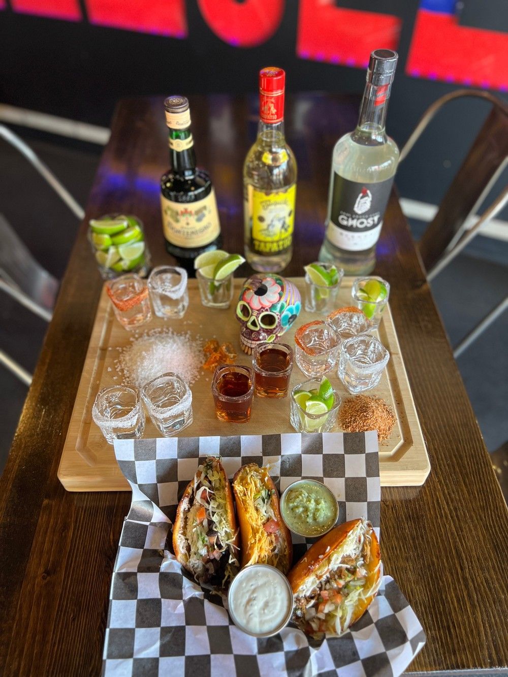 Overhead shot of a tequila flight and Mexican-style tacos on a wooden table — three loaded tacos with sauces and guacamole in a checkered basket, multiple shot glasses with lime wedges, salt and chili rims, colorful decorative skull and three spirit bottles.