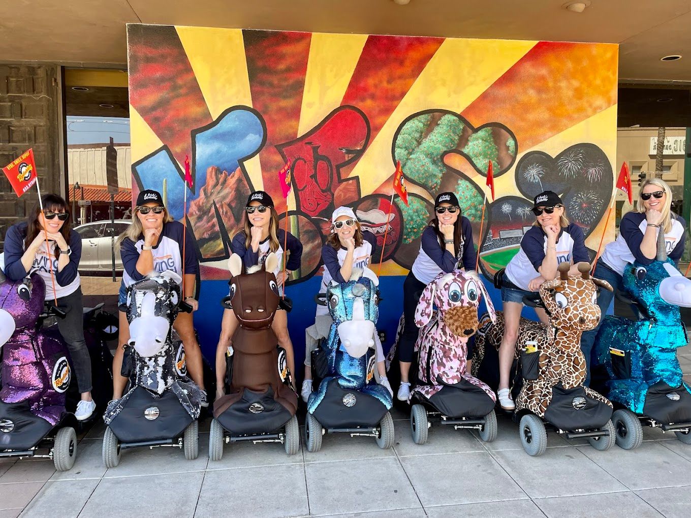 Seven women in matching shirts and caps riding colorful motorized animal scooters (unicorns, horse, giraffe and more) lined up on a sidewalk in front of a vibrant sunburst graffiti mural.