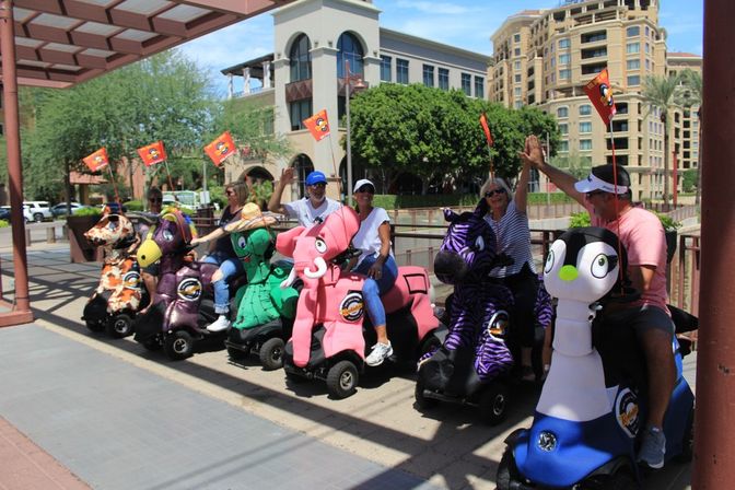 Riders on colorful animal-themed mobility scooters (pink elephant, purple zebra, penguin, green turtle) lined up on a sunny urban waterfront promenade, laughing and high-fiving during an outdoor scooter tour
