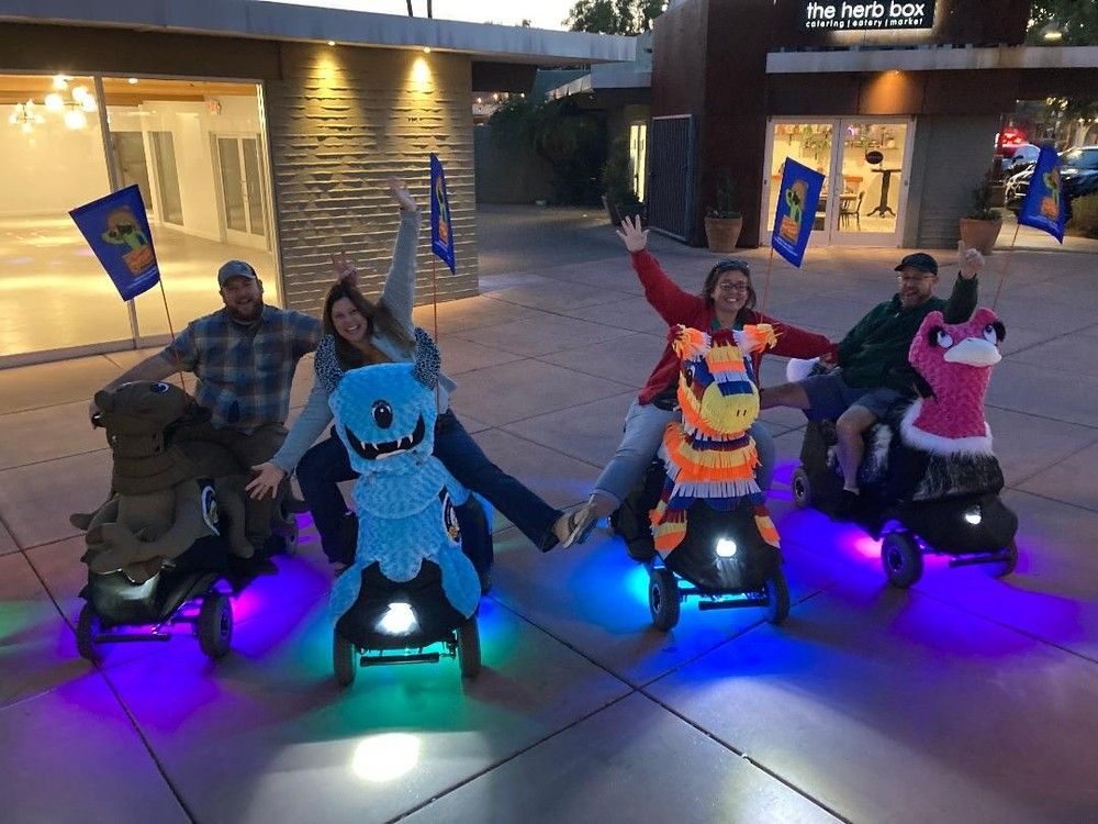 Four people riding novelty animal-themed mobility scooters — brown camel, blue cartoon monster, colorful piñata horse, and pink flamingo — waving and smiling in an outdoor plaza at dusk with purple and blue LED underglow and small flags.
