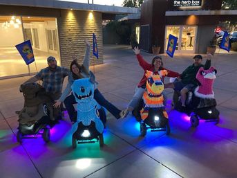Four people riding novelty animal-themed mobility scooters — brown camel, blue cartoon monster, colorful piñata horse, and pink flamingo — waving and smiling in an outdoor plaza at dusk with purple and blue LED underglow and small flags.
