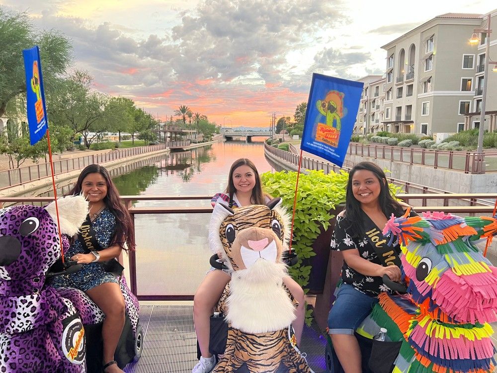 Three smiling people riding colorful animal-themed mobility scooters on a bridge over a calm urban canal at sunset, with palm trees, apartment buildings, festive flags and a vibrant sky reflected in the water.
