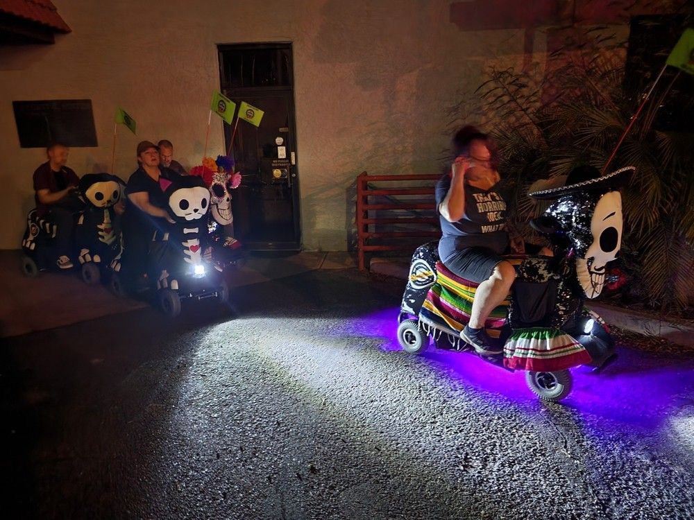 Night street scene of a small parade of decorated mobility scooters with skull and Day of the Dead–style faces, colorful flowers, a sombrero and striped serape, purple underglow lights and small flags, riders cruising past a building wall.