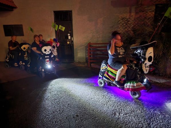Night street scene of a small parade of decorated mobility scooters with skull and Day of the Dead–style faces, colorful flowers, a sombrero and striped serape, purple underglow lights and small flags, riders cruising past a building wall.
