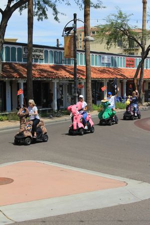 Four adults riding colorful animal-shaped mobility scooters on a sunny downtown shopping street lined with palm trees and tile-roofed storefronts.