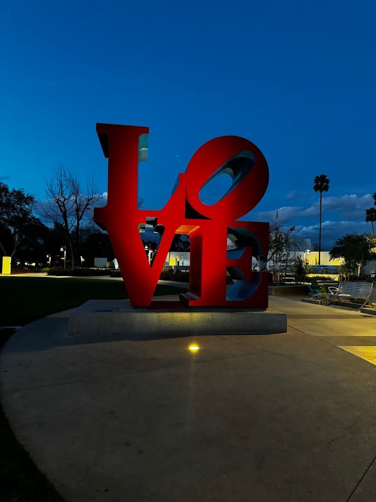 Giant red "LOVE" sculpture glowing at dusk in a lit urban plaza, with palm trees, benches, and a deep blue evening sky