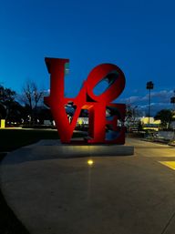 Giant red "LOVE" sculpture glowing at dusk in a lit urban plaza, with palm trees, benches, and a deep blue evening sky