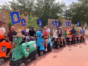 Group of adults riding colorful piñata-style mobility scooters in a sunny outdoor plaza, lined up with small flags and trees in the background
