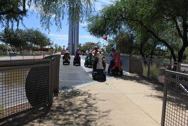 Group of people on decorated mobility scooters with animal-themed covers and orange safety flags riding across a sunny urban park bridge over a canal, surrounded by trees and a paved walkway.