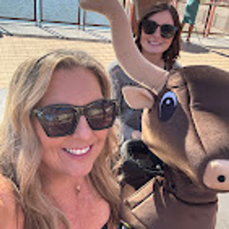 Two smiling women in sunglasses take a selfie with a playful bull statue on a sunny waterfront boardwalk