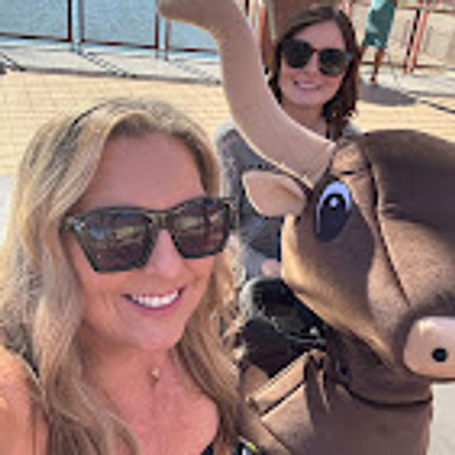 Close-up selfie of two women in sunglasses smiling with a large brown bull mascot on a sunny waterfront pier