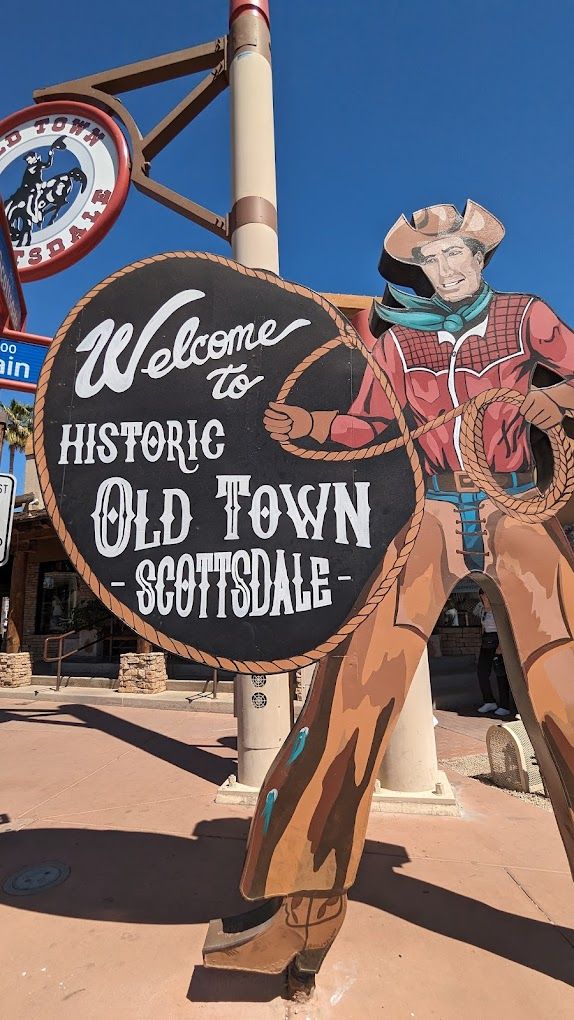 Painted giant cowboy cutout holding a rope and a round sign that reads 'Welcome to Historic Old Town Scottsdale' against a clear blue sky.