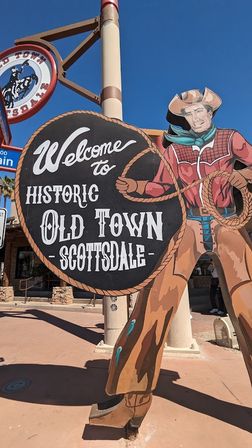 Painted giant cowboy cutout holding a rope and a round sign that reads 'Welcome to Historic Old Town Scottsdale' against a clear blue sky.