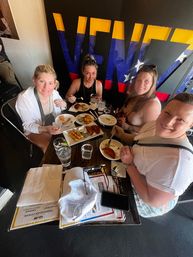 Four friends enjoying Venezuelan-style appetizers—empanadas and fried snacks—with drinks at a small table in front of a bold yellow-blue-red mural.