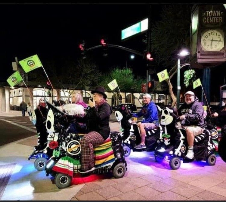 Night downtown scene of a group riding decorated, illuminated mobility scooters with colorful blankets, skull/zebra-themed designs and small green flags beneath a town-center clock