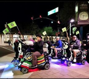 Night downtown scene of a group riding decorated, illuminated mobility scooters with colorful blankets, skull/zebra-themed designs and small green flags beneath a town-center clock