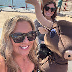 Two women in sunglasses smiling and posing with a playful bull mascot on a sunny waterfront boardwalk.