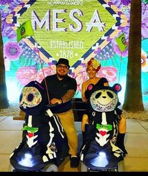 Two adults smiling on decorated Day-of-the-Dead skeleton-panda mobility scooters in front of a colorful MESA mural reading “Established 1878” with large florals — outdoor evening scene.