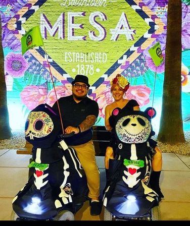 Two adults smiling on decorated Day-of-the-Dead skeleton-panda mobility scooters in front of a colorful MESA mural reading “Established 1878” with large florals — outdoor evening scene.