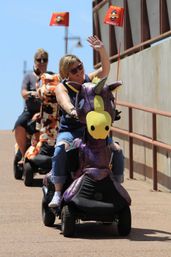 Two people riding decorated mobility scooters on a sunny promenade — front rider waving from a sparkly purple unicorn scooter, followed by a rider on an orange cow-print scooter.