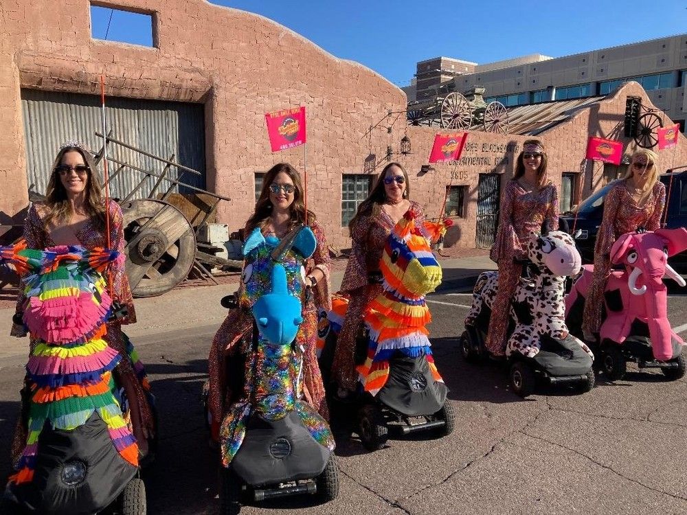 Group of women in shimmering outfits riding mobility scooters dressed as colorful piñata-style animals (rainbow horse, blue unicorn, striped llama, spotted cow, pink elephant) on a sunlit street in front of a southwestern adobe building under a clear blue sky.