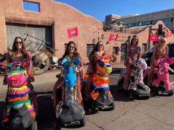 Group of women in shimmering outfits riding mobility scooters dressed as colorful piñata-style animals (rainbow horse, blue unicorn, striped llama, spotted cow, pink elephant) on a sunlit street in front of a southwestern adobe building under a clear blue sky.