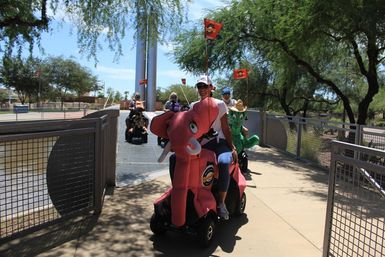 Group of adults riding decorated mobility scooters — a pink elephant and green cactus — across a sunny park bridge with trees and flags
