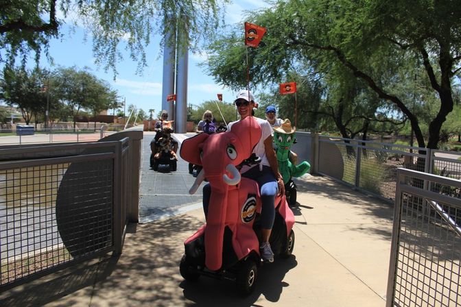 Group of adults riding decorated mobility scooters — a pink elephant and green cactus — across a sunny park bridge with trees and flags