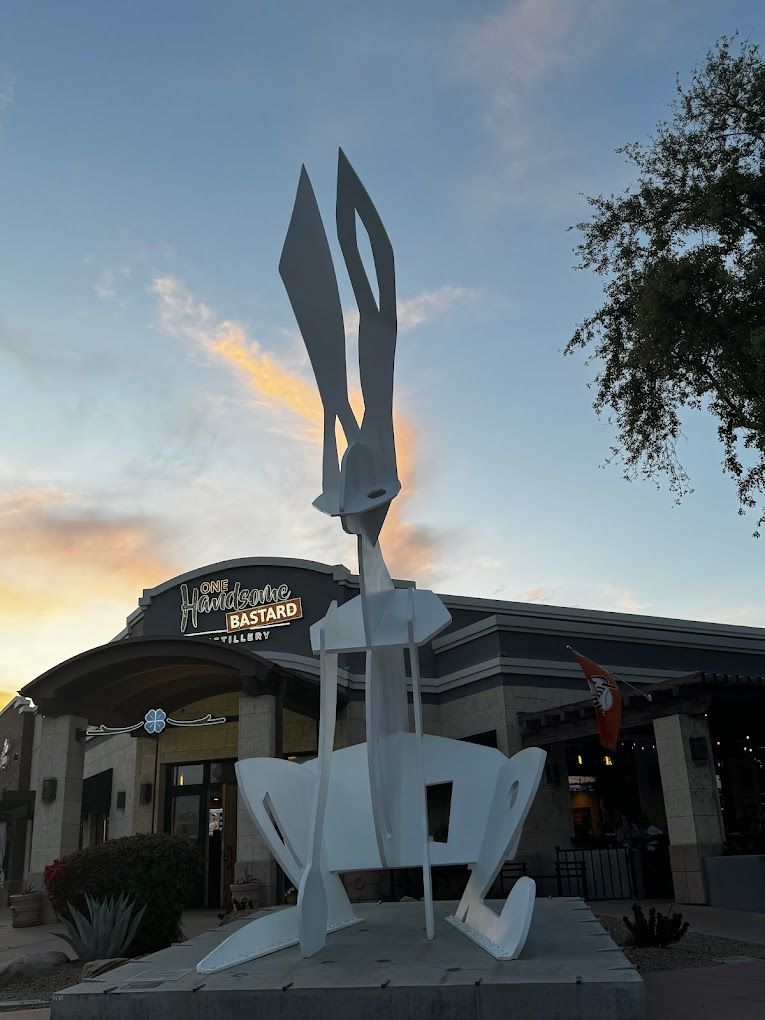 Tall abstract white metal sculpture with blade-like forms rising above a storefront and desert plants in an outdoor plaza at sunset, modern public art piece against a colorful sky.