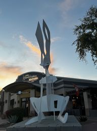 Tall abstract white metal sculpture with blade-like forms rising above a storefront and desert plants in an outdoor plaza at sunset, modern public art piece against a colorful sky.
