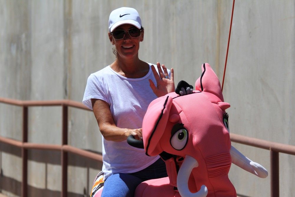 Person in a white cap and sunglasses waving and smiling while riding a bright pink elephant amusement ride along a sunlit concrete walkway.