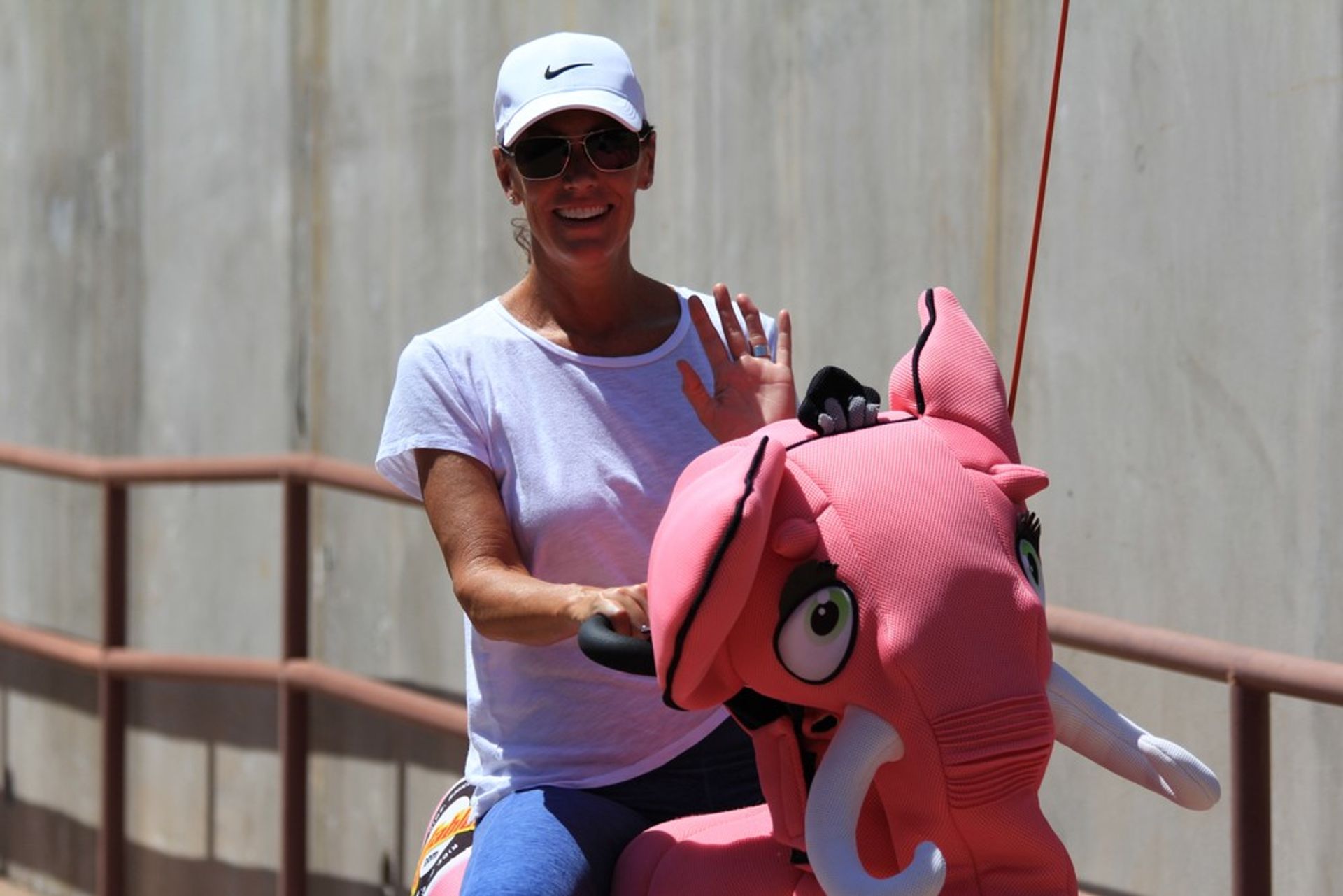 Person in a white cap and sunglasses waving and smiling while riding a bright pink elephant amusement ride along a sunlit concrete walkway.