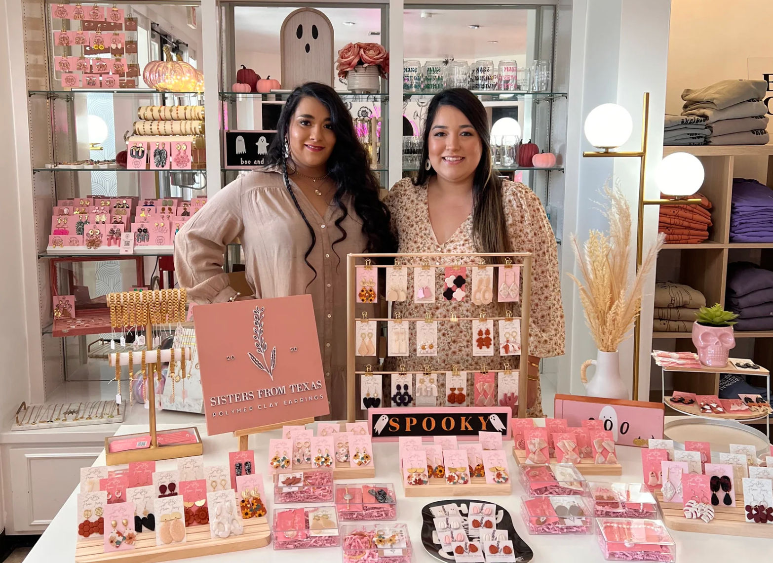 Two smiling vendors at a boutique pop-up standing behind a pink-themed table of handmade polymer clay earrings and jewelry displays, with seasonal pumpkins, shelves of accessories, and a small “spooky” sign in the background.