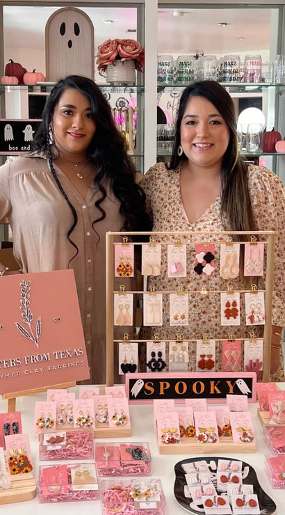 Two smiling vendors at a boutique pop-up standing behind a pink-themed table of handmade polymer clay earrings and jewelry displays, with seasonal pumpkins, shelves of accessories, and a small “spooky” sign in the background.