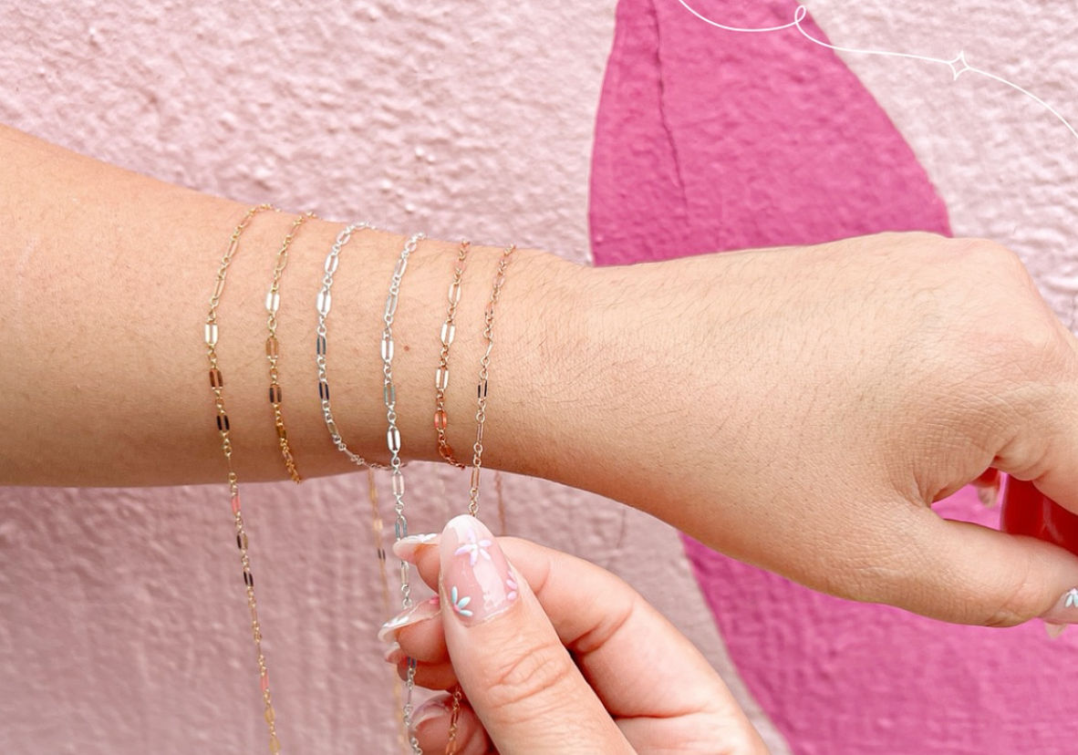 Close-up of a wrist stacked with delicate gold, rose gold and silver chain bracelets against a pink textured wall, another hand with floral nail art adjusting a chain.